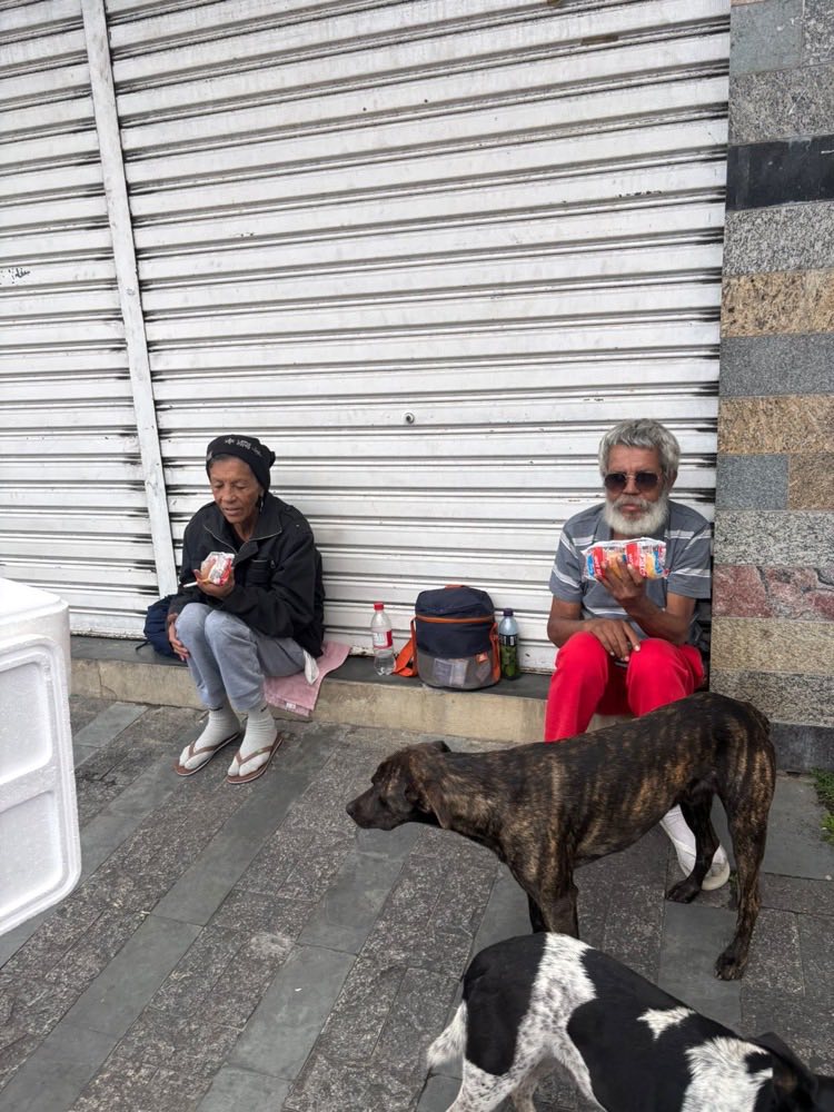 Uma mulher e um homem sentados em frente a uma loja fechada, com lanches nas mãos, e dois cachorros por perto